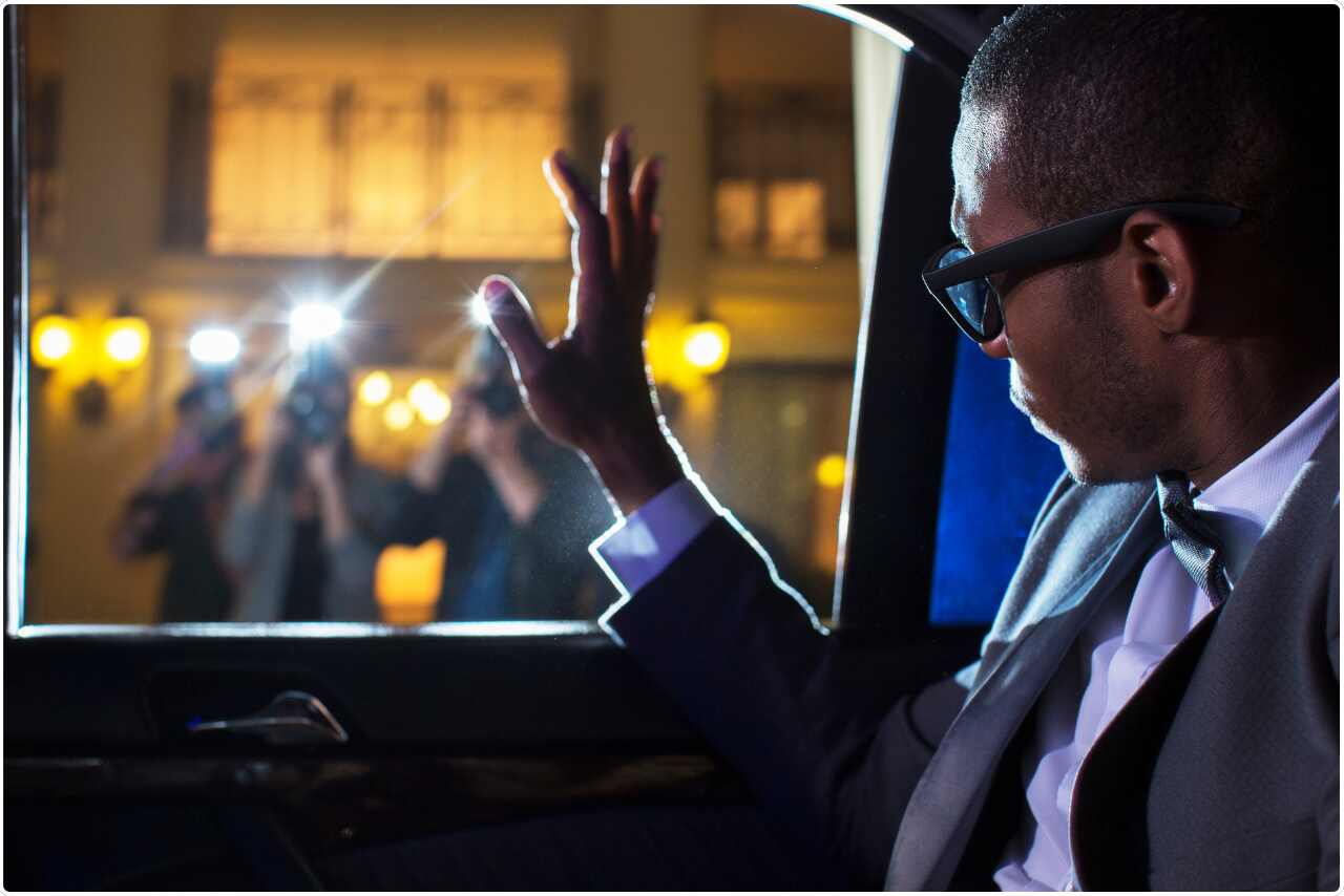 Famous person smiling and waving at photographers through inside car limo  window. 